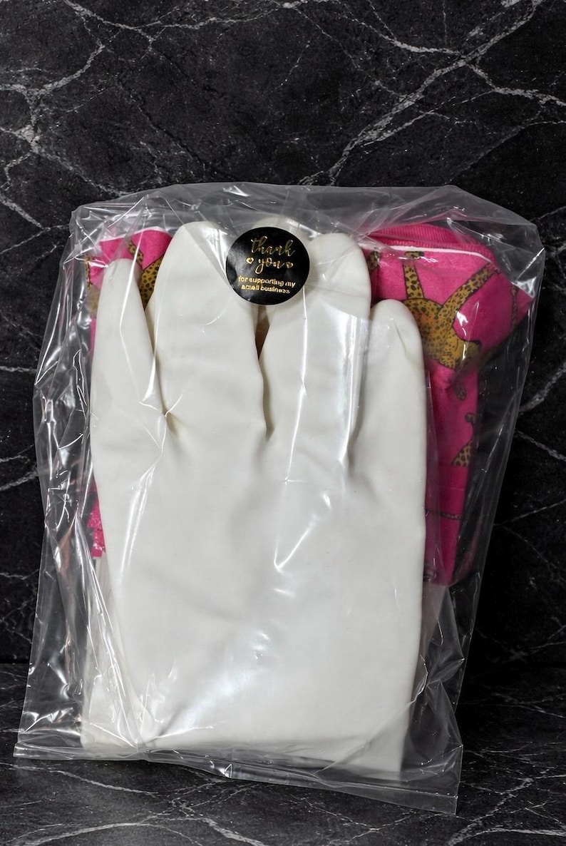 May include: A clear plastic bag containing a white glove and a pink fabric item with a gold pattern. A black sticker with gold text that says "Thank you" is visible on the bag.