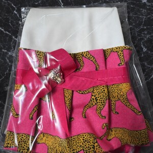 May include: A pink and white dress with a cheetah print skirt. The skirt features a repeating pattern of yellow and black cheetahs on a pink background. A pink ribbon and bow with a decorative jewel accent the waist.