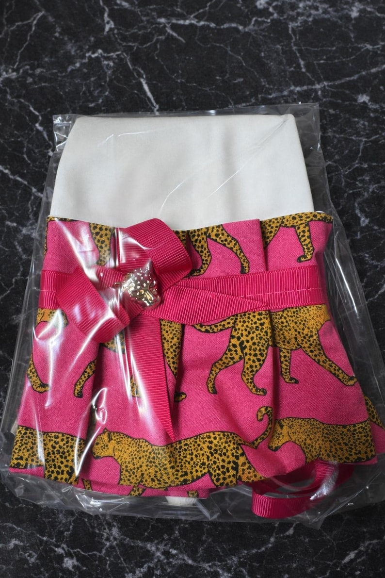 May include: A pink and white dress with a cheetah print skirt. The skirt features a repeating pattern of yellow and black cheetahs on a pink background. A pink ribbon and bow with a decorative jewel accent the waist.