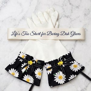 May include: A pair of white dish gloves featuring black ruffled cuffs with a white daisy print. Each cuff is accented with a black ribbon bow and a small yellow bee embellishment. A cream-colored ribbon band across the gloves reads, Life's Too Short for Boring Dish Gloves.