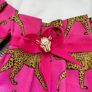 May include: Close-up of a pink fabric with a leopard print in black and gold. A bright pink ribbon is tied in a bow, adorned with a gold-colored animal-shaped embellishment. The fabric is pleated.