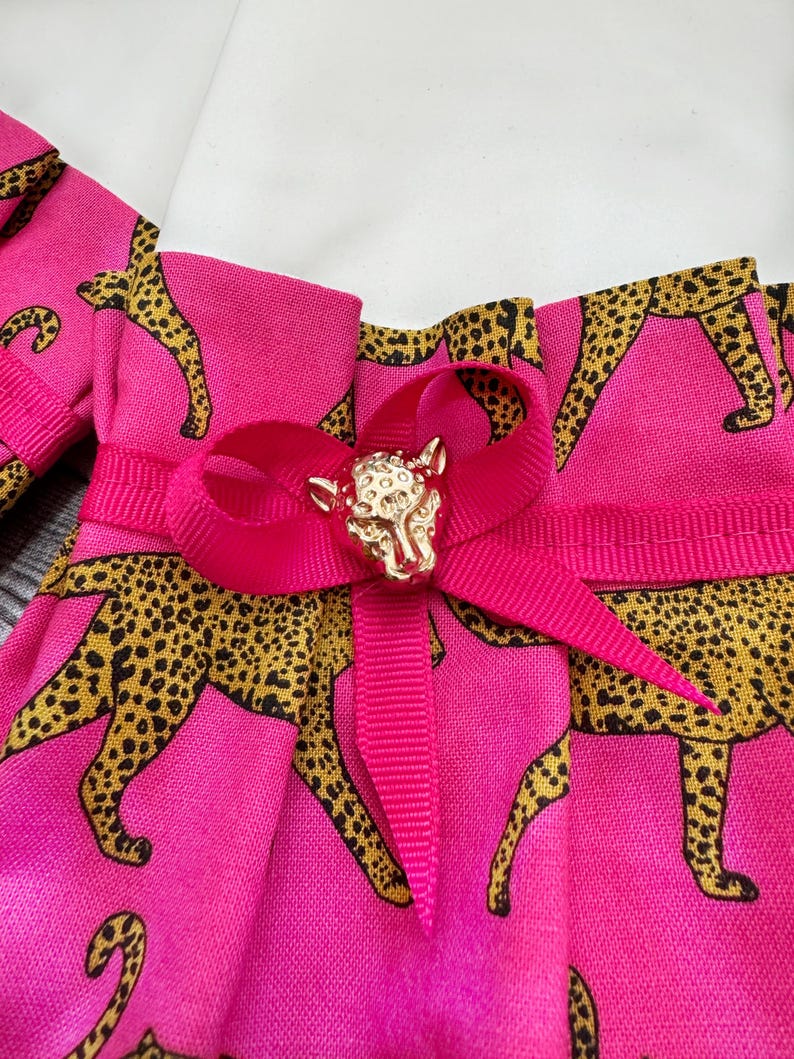 May include: Close-up of a pink fabric with a leopard print in black and gold. A bright pink ribbon is tied in a bow, adorned with a gold-colored animal-shaped embellishment. The fabric is pleated.