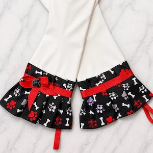 May include: White oven mitts with black and red paw print and bone pattern cuffs. A red ribbon and bow with a paw print accent the cuffs. The mitts are designed for kitchen use.