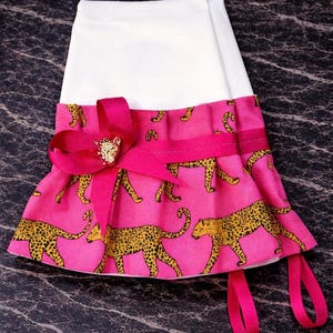 May include: A white and pink pet accessory with a leopard print design. The pink fabric features yellow leopard spots. A bright pink ribbon and bow with a gold-colored embellishment adorn the front. Two pink ribbons hang from the bottom.