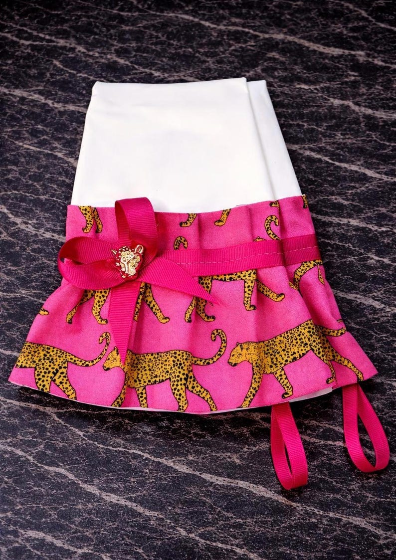 May include: A white and pink pet accessory with a leopard print design. The pink fabric features yellow leopard spots. A bright pink ribbon and bow with a gold-colored embellishment adorn the front. Two pink ribbons hang from the bottom.