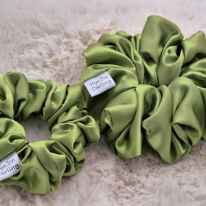 Pode incluir: Duas scrunchies de cetim verde-oliva. Uma é menor e a outra maior. Cada scrunchie tem uma etiqueta branca com a inscrição "HyeJin Darling". As scrunchies estão sobre uma superfície fofa e clara.