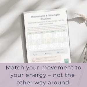 May include: A white planner titled "Movement & Strength Planner" with a grid of boxes and the text "Match your movement to your energy - not the other way around." The planner is on a white surface with a shadow.