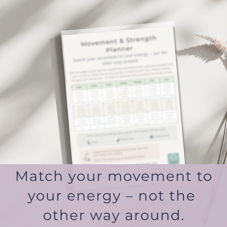 May include: A white planner titled "Movement & Strength Planner" with a grid of boxes and the text "Match your movement to your energy - not the other way around." The planner is on a white surface with a shadow.