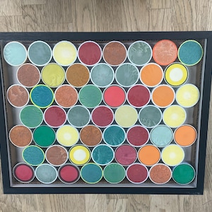 Puede incluir: Una obra de arte enmarcada con una cuadrícula de objetos circulares coloridos. Los círculos muestran una variedad de colores, incluyendo rojo, verde, naranja, amarillo y marrón. La obra está encerrada en un marco negro.
