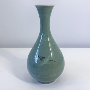 May include: A celadon-colored ceramic vase with a slender neck and rounded body. The vase features a delicate illustration of a bird in flight and subtle etched lines. The vase is approximately 7 inches tall.