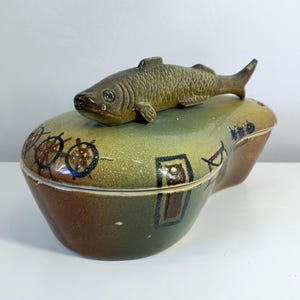 May include: A ceramic trinket box with a fish-shaped lid. The box is oval-shaped with a green, brown, and tan color scheme. The lid features a detailed fish sculpture. Decorative nautical-themed accents are painted on the box.