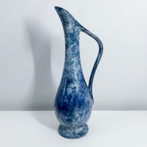 May include: A tall, blue and white ceramic pitcher with a curved handle. The pitcher has a speckled pattern and a flared spout. The base is round and slightly raised. The pitcher is likely used for serving water or other beverages.