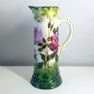 May include: Tall, antique ceramic pitcher with a cream-colored background. The pitcher is decorated with pink and purple roses and green leaves. The handle is cream-colored with gold accents. The base and top rim are green.