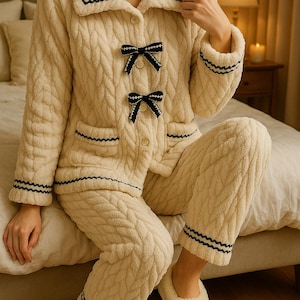 Könnte beinhalten: Cremefarbenes Pyjama-Set mit strukturiertem Zopfmuster. Die Jacke hat schwarze Schleifenakzente und -besätze sowie Taschen. Die Hose hat passende Besätze an den Knöcheln. Das Set enthält passende Hausschuhe.