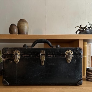 May include: A black leather vintage toolbox with brass hardware and a handle. The toolbox is sitting on a wooden shelf with other decorative items.