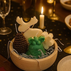 May include: A wooden bowl containing decorative candles shaped like a reindeer, pinecone, and a green figure. The candles are white, brown, and green. The bowl is on a black tablecloth with gold stars, near a lit candle and a wine glass.