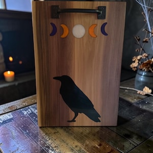 May include: A rectangular wooden serving tray featuring a black raven silhouette and a moon phase graphic in purple, orange, and white. The tray includes a dark metal handle mounted at the top centre, showcasing natural wood grain patterns.