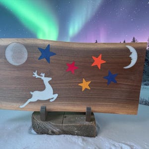 May include: A rectangular wooden board with a live edge featuring inlaid resin designs. The inlay includes a white leaping deer, a full moon, a crescent moon with a face, and five stars in blue, red, orange, and yellow colours on the wood surface.