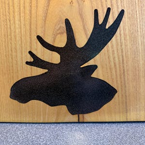 May include: A light brown wooden board featuring a black, textured silhouette of a moose head with prominent antlers. The wood grain is visible throughout the background, providing a natural contrast to the dark, matte finish of the animal shape.