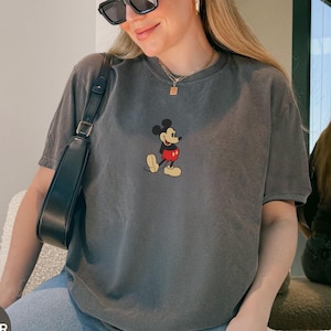 May include: A gray t-shirt featuring a classic Mickey Mouse graphic. The short-sleeved shirt has a crew neck and a slightly faded appearance. The graphic shows Mickey Mouse in his iconic pose, with red shorts and yellow shoes.