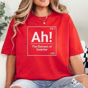 May include: A red t-shirt with the text "Ah! The Element of Surprise" in a white box. The shirt has short sleeves and a relaxed fit. The text includes the numbers 29.7 and 230.