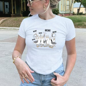 May include: White t-shirt featuring a cartoon illustration of seagulls with the word "MOM!" above each bird. The seagulls are in various poses, with gray and white coloring. The shirt has short sleeves and a crew neck.