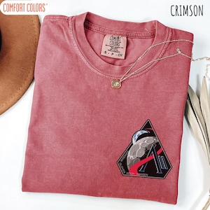 May include: Crimson Comfort Colors t-shirt with a crew neck and a small patch featuring a moon and the text "WISEMAN GLOVER KOCH HANSEN". A silver necklace with a pendant is draped across the shirt.