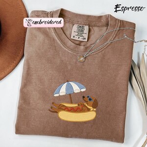 May include: A brown crewneck t-shirt with an embroidered graphic of a dachshund wearing heart-shaped sunglasses, resting inside a hot dog bun under a blue and white striped beach umbrella. The shirt features a Comfort Colors brand label.