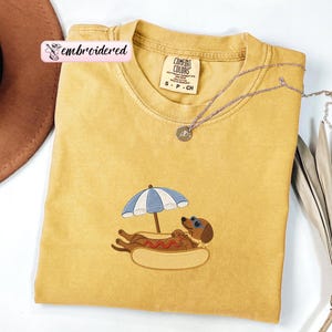 May include: A mustard yellow short-sleeve t-shirt featuring an embroidered graphic of a brown dachshund wearing blue sunglasses, resting inside a hot dog bun under a blue and white striped beach umbrella. The shirt has a Comfort Colors label.