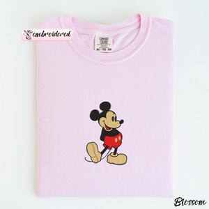 Peut inclure: Un t-shirt rose clair &agrave; col rond et manches courtes. Le t-shirt pr&eacute;sente une image brod&eacute;e de Mickey Mouse dans sa tenue classique : short noir, pantalon rouge et chaussures jaunes. Le t-shirt est pli&eacute; proprement.