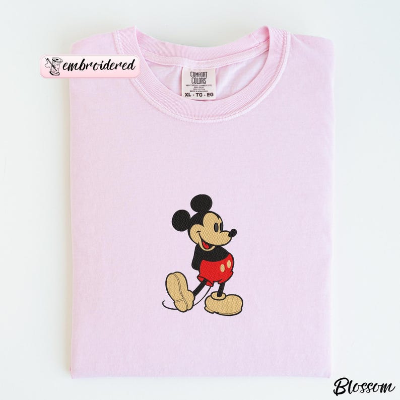 Peut inclure: Un t-shirt rose clair &agrave; col rond et manches courtes. Le t-shirt pr&eacute;sente une image brod&eacute;e de Mickey Mouse dans sa tenue classique : short noir, pantalon rouge et chaussures jaunes. Le t-shirt est pli&eacute; proprement.