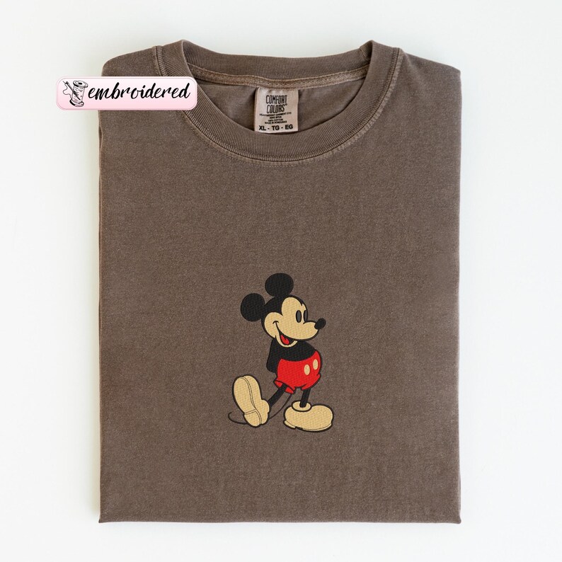Peut inclure: Un t-shirt marron avec un motif brod&eacute; de Mickey Mouse. Le personnage de dessin anim&eacute; est repr&eacute;sent&eacute; avec un short rouge, une chemise noire et des chaussures jaunes. Le t-shirt a un col rond et une &eacute;tiquette qui dit "Comfort Colors". Le mot "brod&eacute;" est sur une &eacute;tiquette rose.
