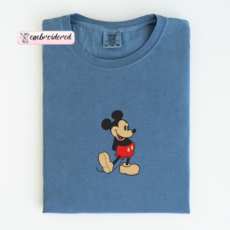 Peut inclure: T-shirt bleu avec une image brod&eacute;e de Mickey Mouse. Le personnage de dessin anim&eacute; porte un short rouge, des chaussures jaunes et une veste noire. Le t-shirt porte le texte "embroidered" sur une &eacute;tiquette rose.