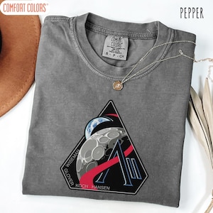 May include: Pepper-colored Comfort Colors t-shirt with a graphic of a moon, Earth, and the letter "A" inside a black triangle. The names "Wiseman", "Glover", "Koch", and "Hansen" are printed on the graphic. A silver necklace with a pendant is on the shirt.