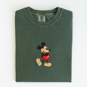 Peut inclure: T-shirt vert fonc&eacute; avec un motif brod&eacute; de Mickey Mouse. Le t-shirt a un col rond et des manches courtes. Le motif Mickey Mouse comprend des couleurs noir, rouge et beige.