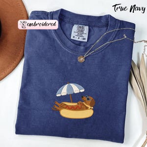 May include: A True Navy short-sleeve t-shirt featuring an embroidered graphic of a brown dachshund wearing blue sunglasses, resting inside a hot dog bun under a blue and white striped beach umbrella. The shirt has a Comfort Colors brand label.