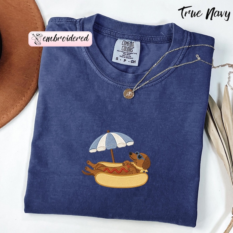 May include: A True Navy short-sleeve t-shirt featuring an embroidered graphic of a brown dachshund wearing blue sunglasses, resting inside a hot dog bun under a blue and white striped beach umbrella. The shirt has a Comfort Colors brand label.