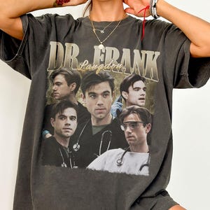 Puede incluir: Camiseta gris oscuro con la inscripción "DR. FRANK Langdon" y un collage de imágenes. La camiseta tiene un aspecto vintage. El diseño presenta múltiples retratos de un hombre con estetoscopio.