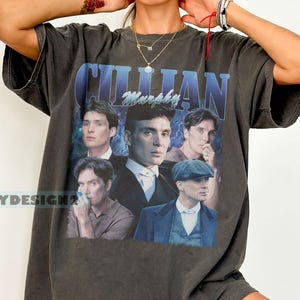 May include: Dark gray t-shirt featuring a collage of images of Cillian Murphy with the name "Cillian Murphy" in blue text. The shirt has a vintage, worn look. The brand name "LEAPYDESIGN2" is printed on the left side of the shirt.