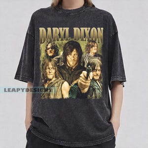 Puede incluir: Camiseta gris oscuro con un gráfico desgastado de Daryl Dixon de The Walking Dead. El diseño incluye el nombre "DARYL DIXON" en letras doradas sobre un collage de imágenes del personaje. La camiseta tiene un aspecto vintage y desgastado.