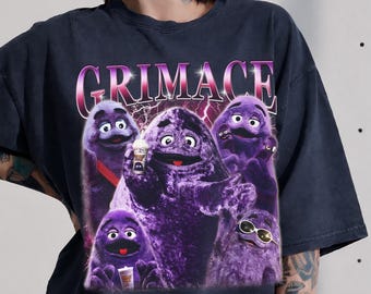 May include: A charcoal gray short-sleeve t-shirt featuring a graphic print of multiple purple Grimace characters. The word GRIMACE is printed in large, stylized pink letters at the top. The design includes lightning effects and a small drink cup.