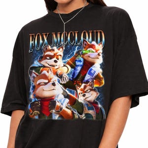 Może przedstawiać: Czarny t-shirt z nadrukiem graficznym przedstawiającym postać Foxa McClouda w czterech różnych pozach. Napis "FOX MCCLOUD" jest wydrukowany pogrubioną czcionką w stylu metalicznym u góry, na ciemnoniebieskim tle z efektami błyskawic.