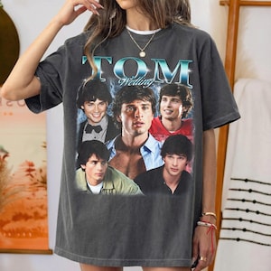 May include: A charcoal gray short-sleeve t-shirt featuring a graphic print of actor Tom Welling. The design includes the text "TOM Welling" in a metallic blue font above four distinct photographic portraits of the actor in various outfits.