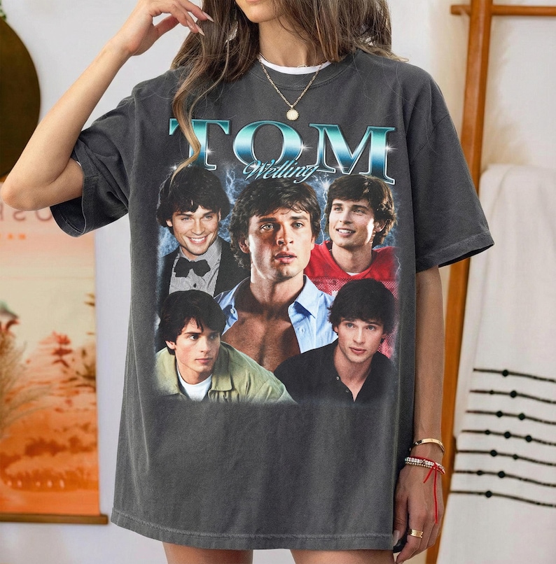 May include: A charcoal gray short-sleeve t-shirt featuring a graphic print of actor Tom Welling. The design includes the text "TOM Welling" in a metallic blue font above four distinct photographic portraits of the actor in various outfits.