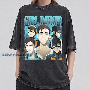 May include: Dark gray t-shirt featuring a graphic of five cartoon figures with the text "GIRL DINNER" in blue. The figures are in various poses, with a blue and white background. The shirt has a vintage, distressed look.