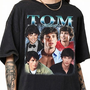 May include: A black short-sleeve t-shirt featuring a graphic print of actor Tom Welling. The design includes the text "TOM Welling" in a metallic blue font at the top, surrounded by five different photographic portraits of the actor in various outfits.