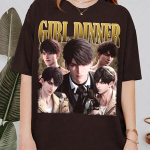 Può includere: T-shirt nero con grafica dorata con la scritta "GIRL DINNER". La grafica include più immagini di un uomo con i capelli scuri e un abito nero.