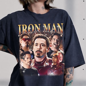 May include: A charcoal gray t-shirt featuring a large graphic print of Iron Man. The design includes the text IRON MAN in a bold yellow font above multiple images of the character in various poses and expressions, including one wearing the helmet.
