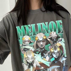 Peut inclure: T-shirt gris avec le mot "MELINOE" en grandes lettres vertes. Le t-shirt présente un graphisme de plusieurs femmes avec des bandeaux dorés et des yeux rouges, sur fond vert. Le design a un thème de bande dessinée ou fantastique.
