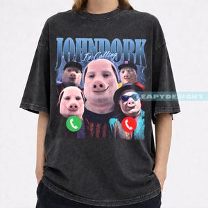 May include: Dark gray t-shirt with a graphic featuring a group of pig-faced people with the text "JOHNDORK Is Calling". The design includes a green and red phone icon.
